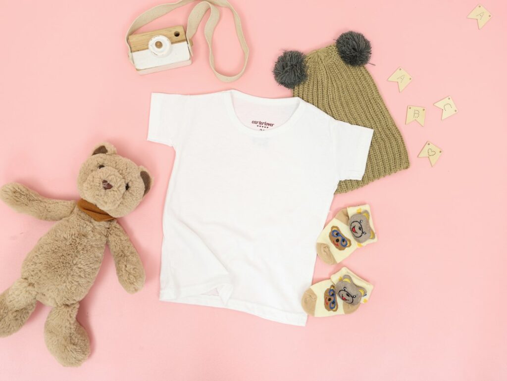 Baby clothes, a knitted hat, socks and a teddy bear arranged on a pink background.