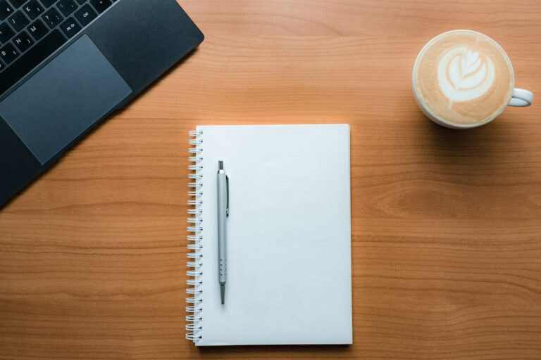 Blank spiral notebook, pen, coffee, and laptop on a wooden table.