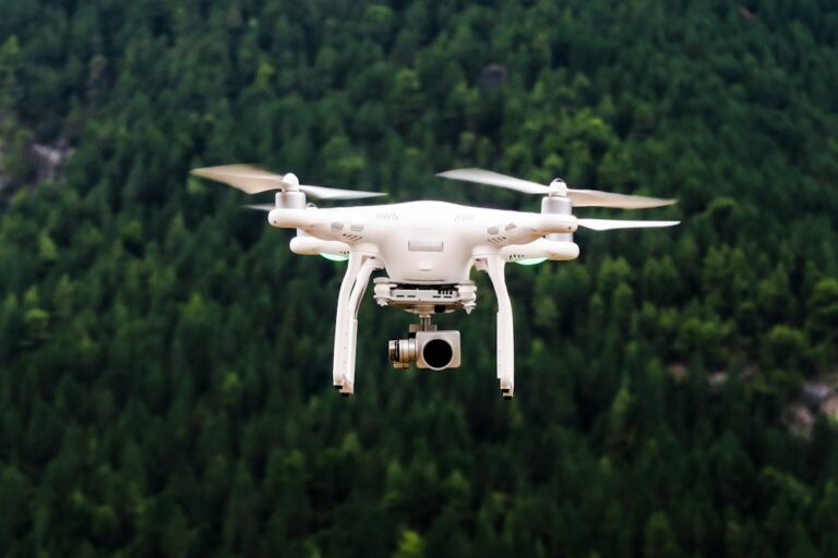 A white quadcopter with a camera flying in front of a wooded hillside.