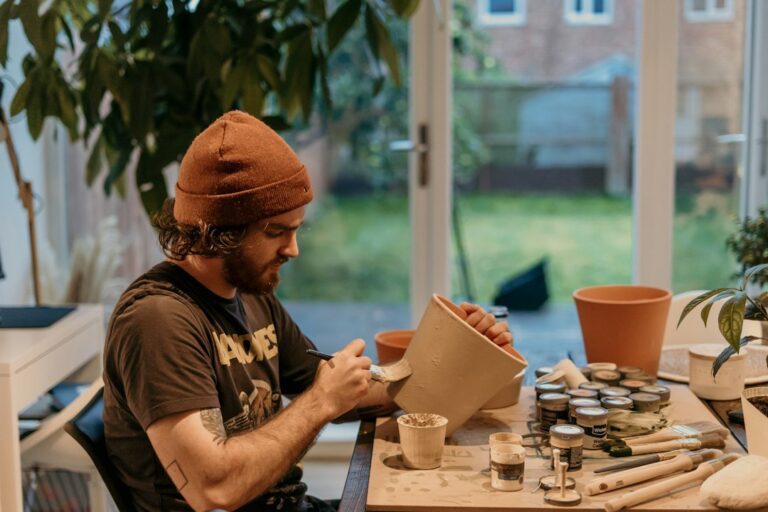 A craftsperson painting a plant pot at a table with brushes and paint containers.