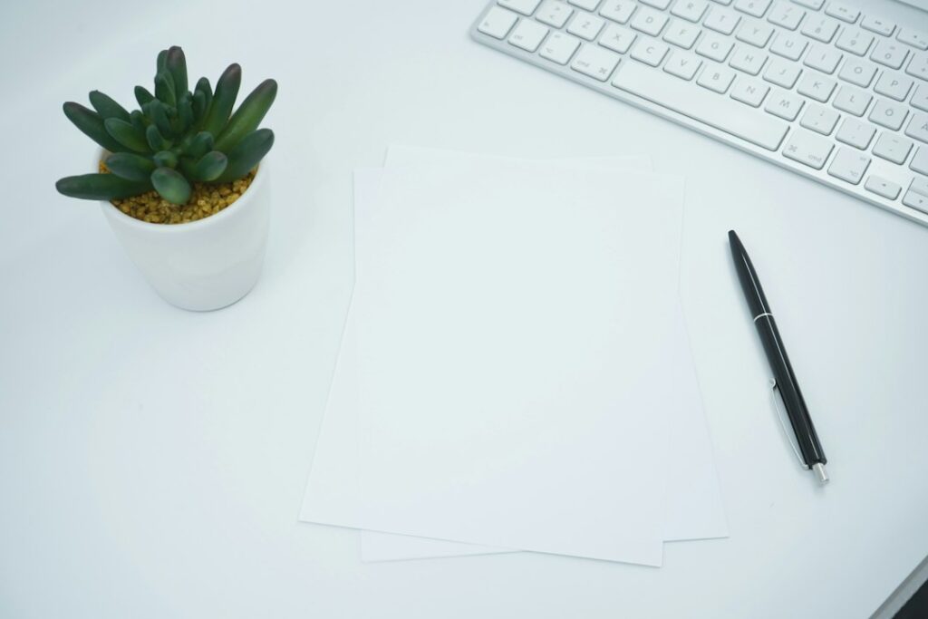 Blank sheets of paper and a black pen on a desk beside a keyboard and small plant.