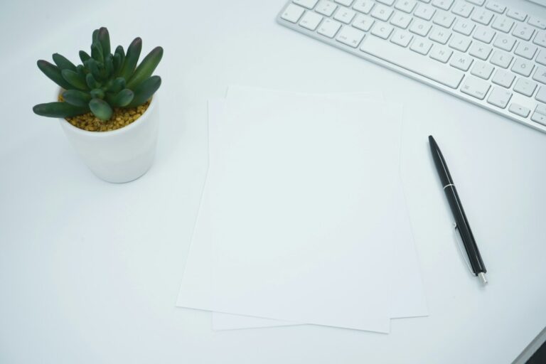 Blank sheets of paper and a black pen on a desk beside a keyboard and small plant.