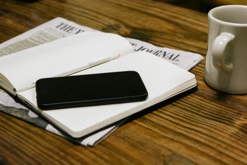 Open notebook, phone, pen, and coffee mug resting on a newspaper.