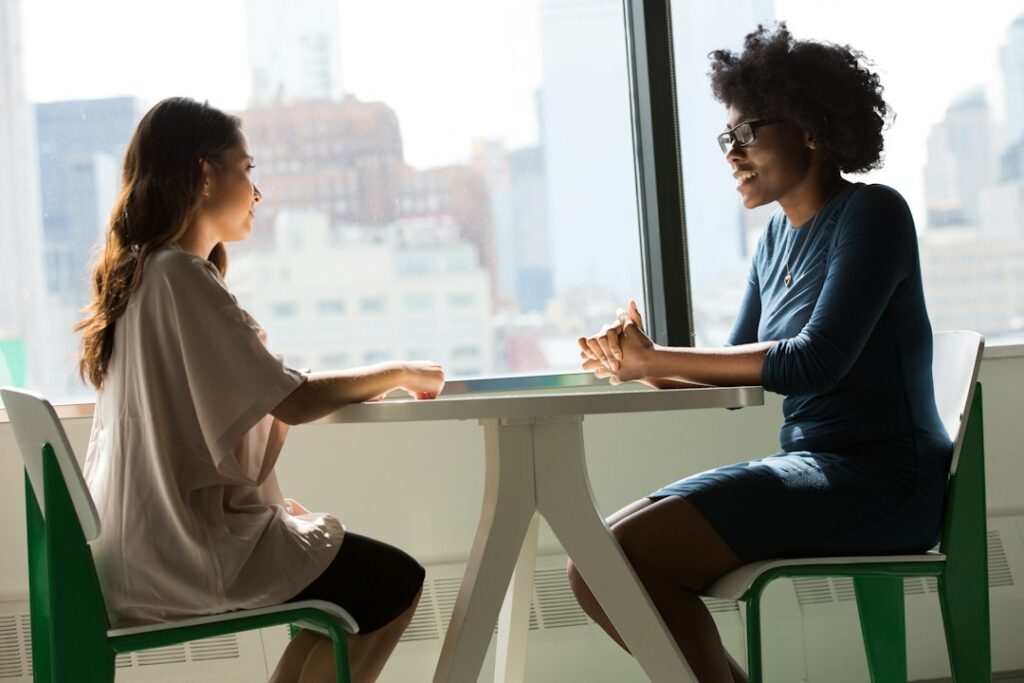 Two women sitting across a table and talking beside a window.