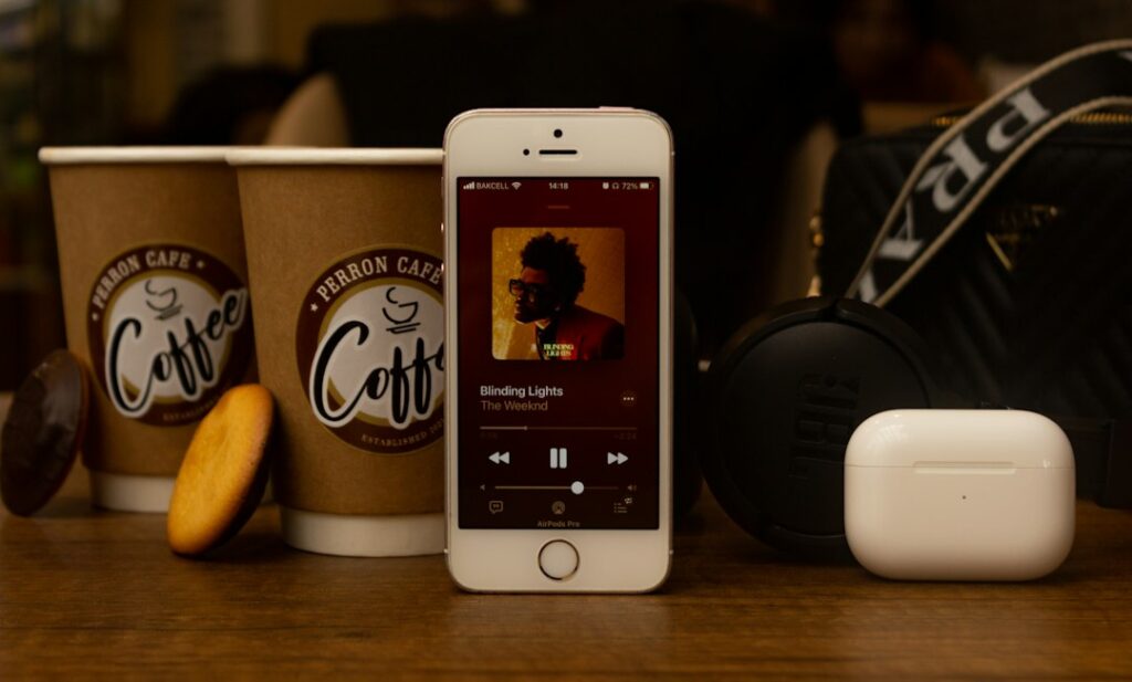 A phone showing a music player beside headphones and takeaway coffee cups