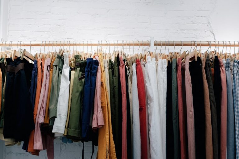 Trousers in many colors hanging from wooden hangers against a white brick wall.