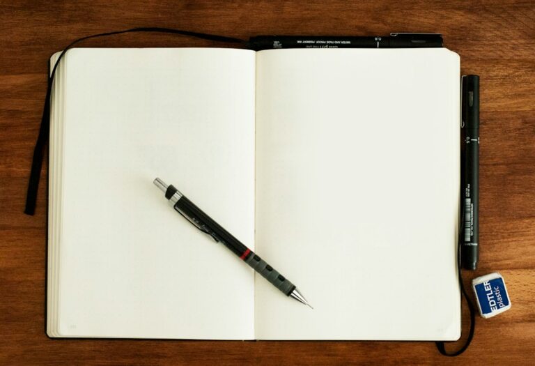 Open blank notebook with a pencil, pens and eraser on a wooden desk.