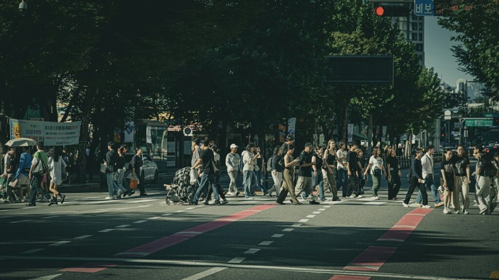 Pedestrians crossing a broad city street beneath trees.