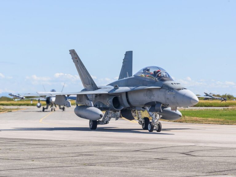 A military jet taxiing on a paved airfield with another aircraft behind it.