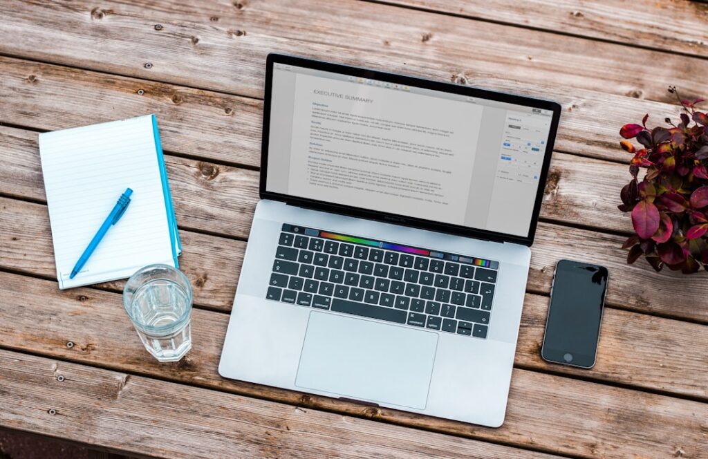 Laptop displaying a document beside a notebook and phone on a wooden table.