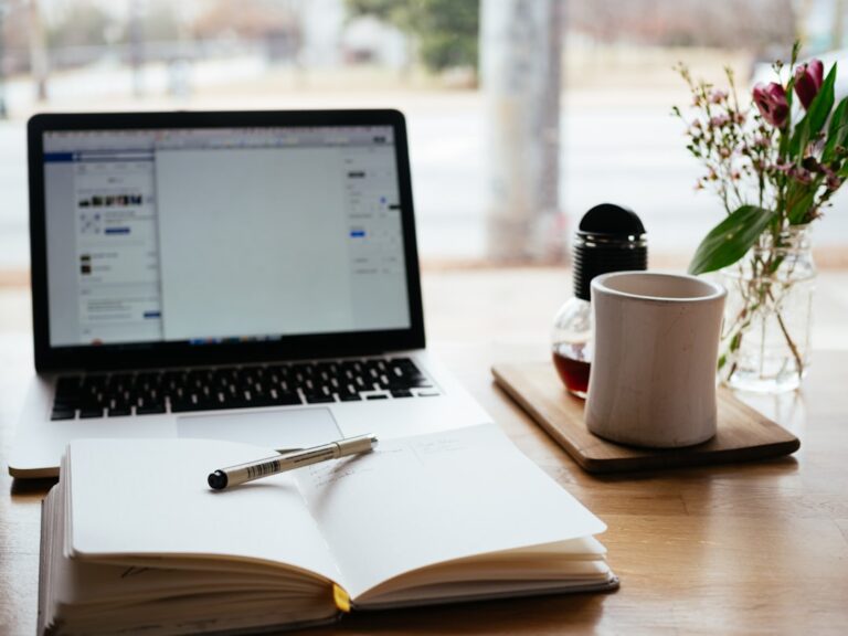 An open notebook and pen beside a laptop, mug, and flowers on a desk.
