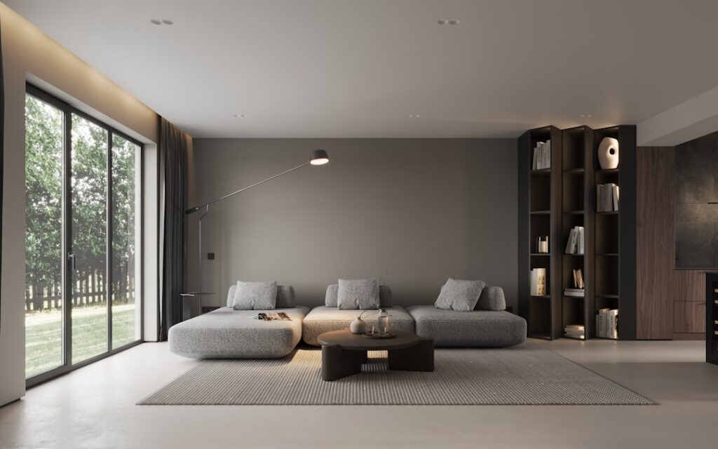 A gray sofa, low coffee table, and shelving in a living room beside large windows.