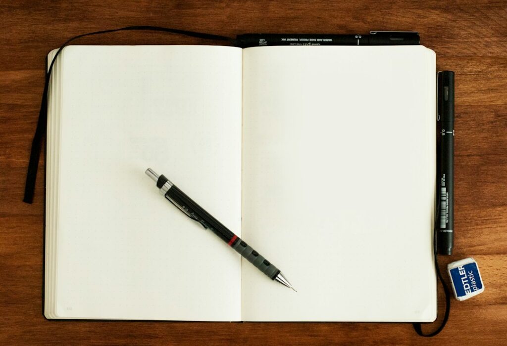 An open blank notebook with pens, a mechanical pencil and an eraser on a wooden desk.