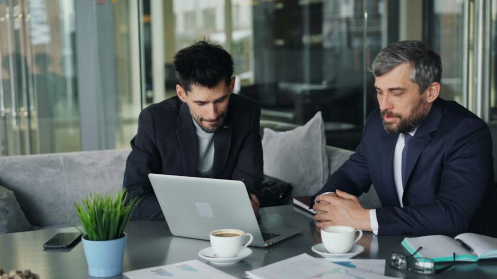 Two men reviewing a laptop together at a table with notebooks.