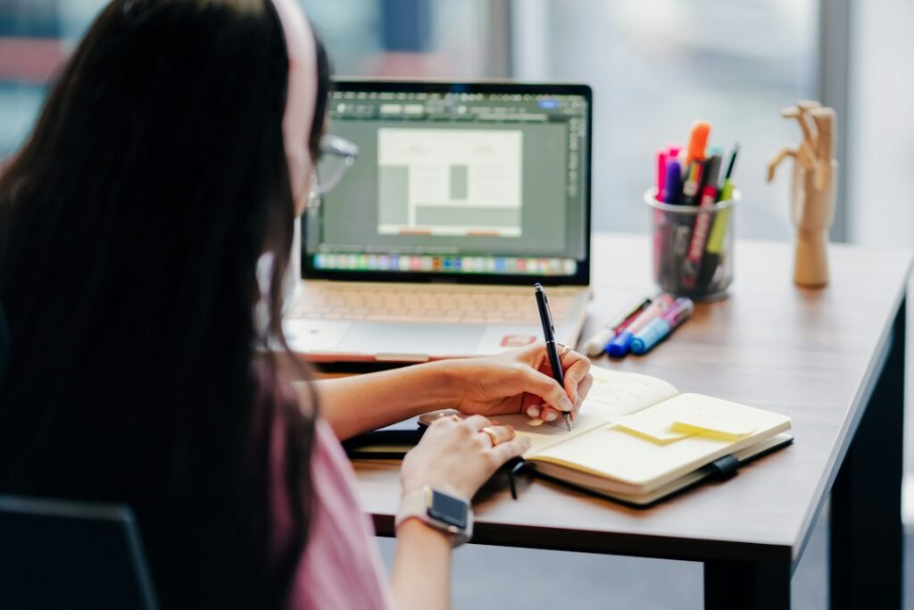 A person writing in a notebook beside a laptop at a home workspace.