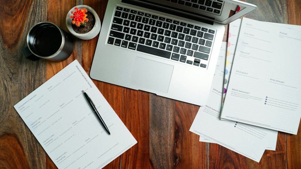 Laptop, printed worksheets, pen, and cup on a wooden desk