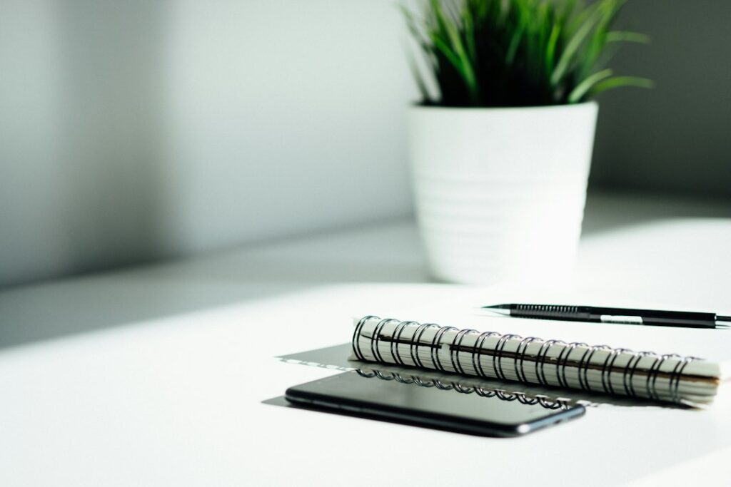 A notebook and pen beside a phone and a potted plant