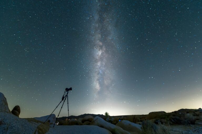 A camera on a tripod beneath a star-filled night sky over rocky ground