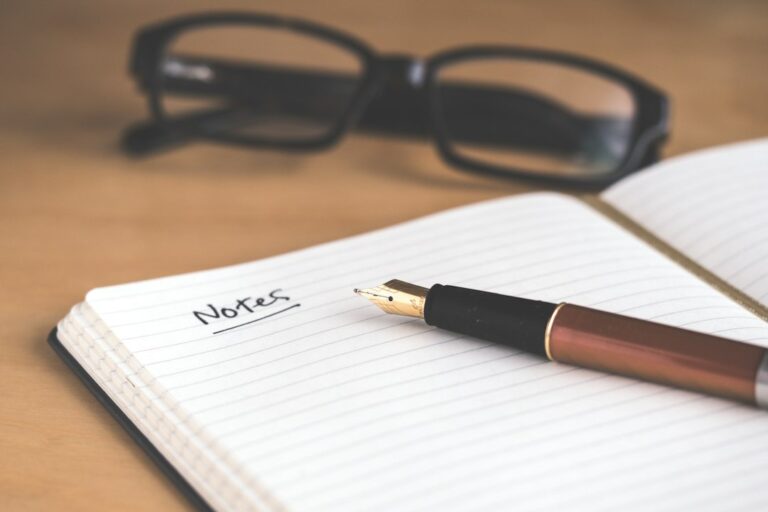 A fountain pen on a lined notebook with reading glasses in the background.