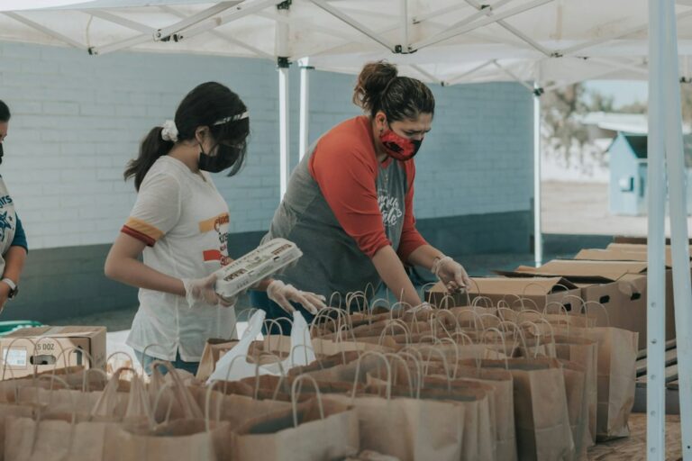 Volunteers wearing face masks and gloves packing food into paper bags under a canopy