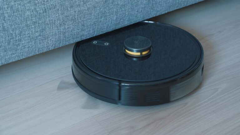 A black circular robot vacuum on a wooden floor beside a sofa.