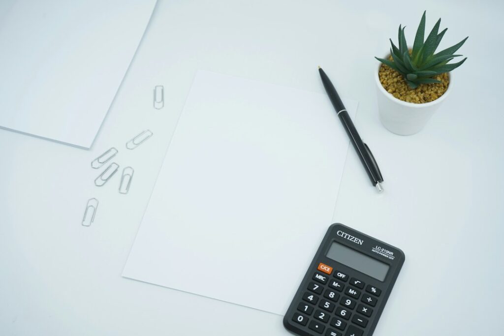 A calculator, pen, blank paper, and small plant arranged on a desk.