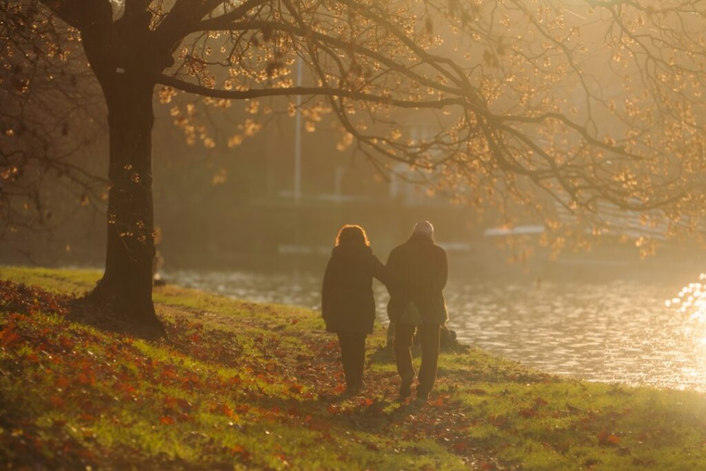 Two people walking beside a river under trees in warm evening light.